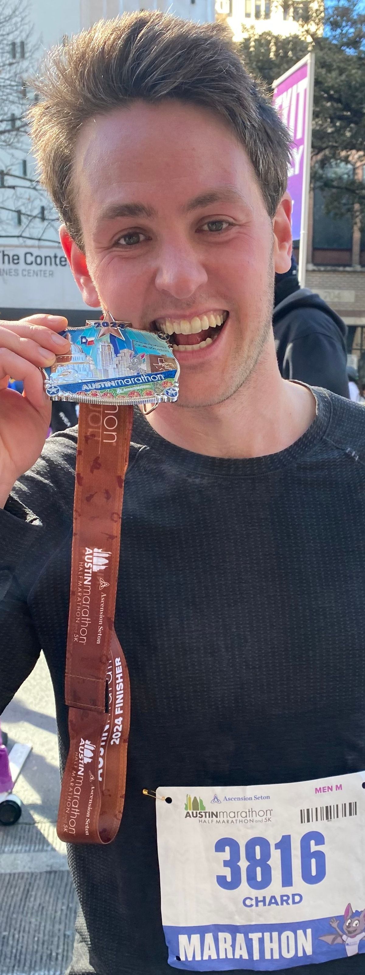 Richard Joseph Spinelli holding his finisher medal up to the camera after the 2024 Austin Marathon, bib number 3816 pinned to his shirt